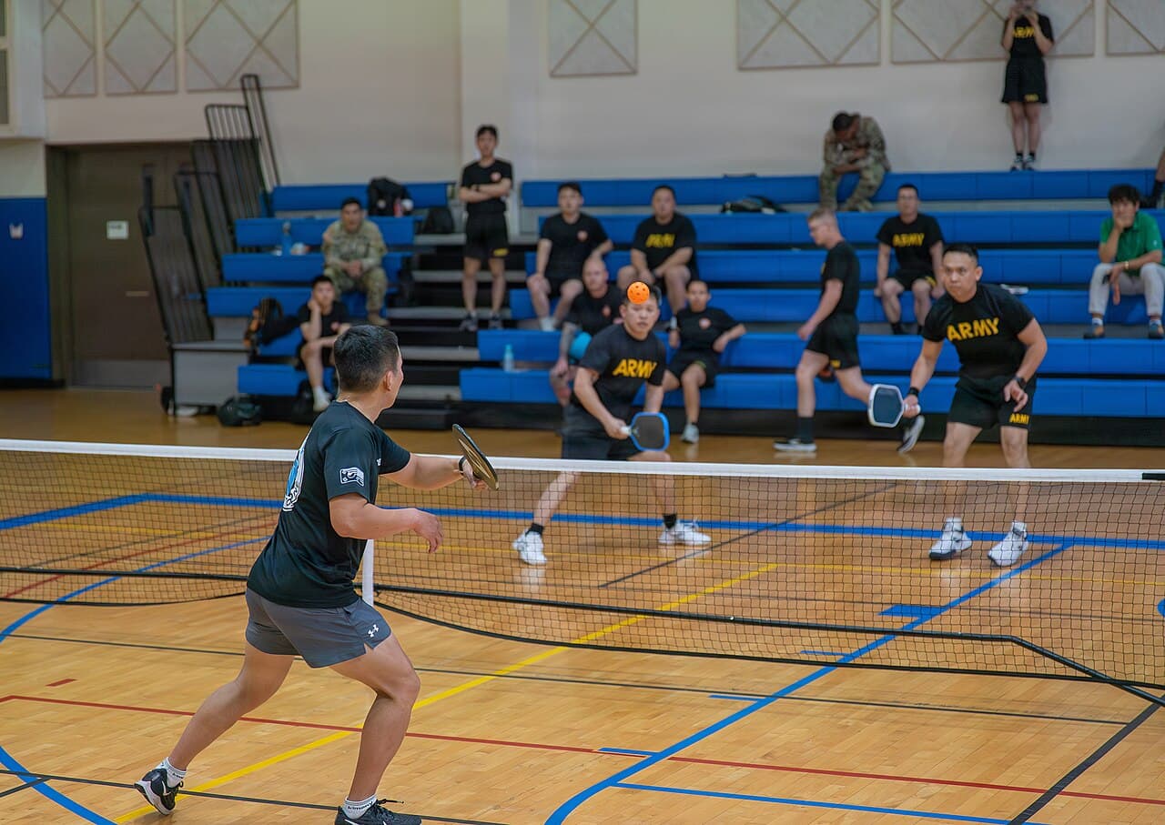 Doubles match on an indoor gym court during an organized pickleball tournament, with teammates and spectators on the bleachers